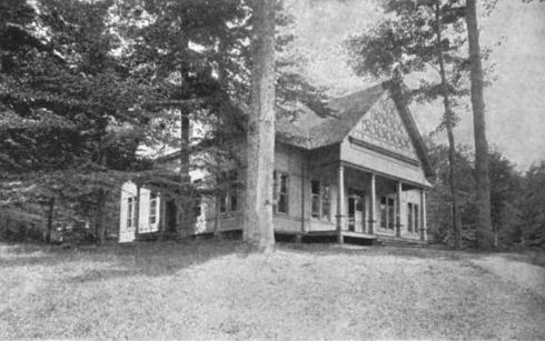 Black and white photo of a building with aa large front porch and gingerbread trim, set among tall trees.