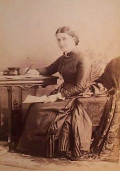 Photograph of Isabella Alden from about 1898. She is seated at a table. In her lap she holds a piece of paper. Her right hand holds a pen poised above a piece of  paper on the table. 
