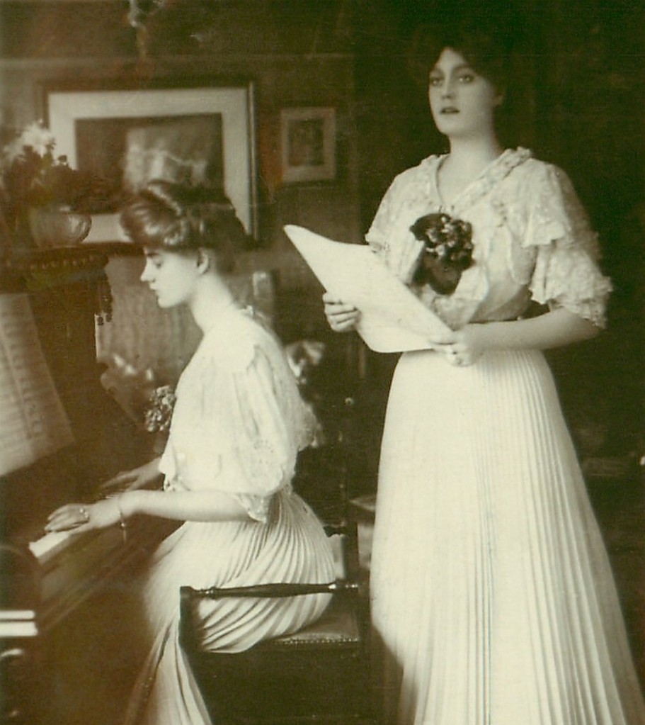 A young woman stands holding sheet music and singing. Beside her another young woman is seated, playing a piano.