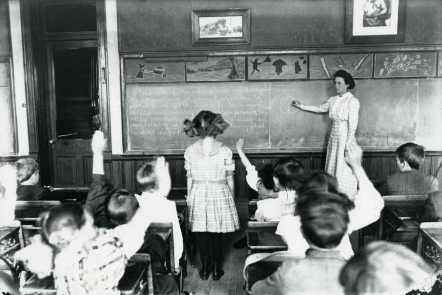 Photo dated about 1910. A teacher stands at the classroom blackboard where words and shapes are drawn, and points to one section of the blackboard. One little girl stands facing the teacher (her back to the camera) to answer the teacher's question. All other students sit at desks with their backs to the camera. Some students have their hands raised to answer the question.