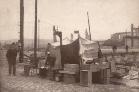A man standing beside his make-shit home made of piled crates and sheets.