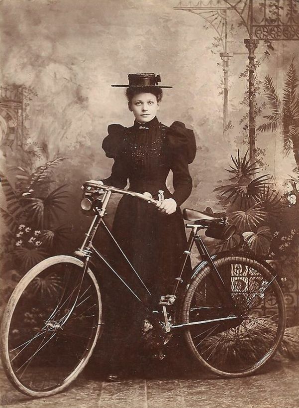 Studio photograph from late 1890s of young woman posed beside her bicycle. She is wearing a long dark dress with long sleeves and a high collar, and a bonnet. 