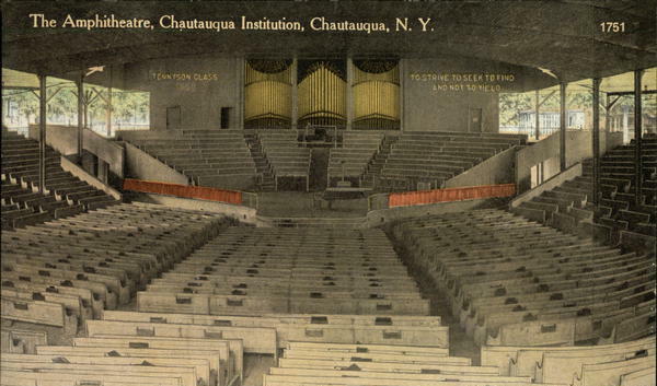 Photo of the inside of the amphitheatre, a large wood structure with a stage at one end with about 12 rows of raised seating behind. The floor and 3 remaining sides of the theatre are filled with rows of wooden benches.  