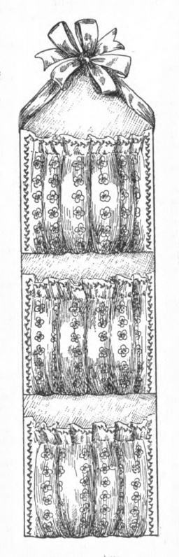 Drawing of a wall pocket made of a long board or cardboard. At one end is a ribbon so it can be hung vertically on the wall. Spaced evenly down the board are three fabric pockets decorated with different trims.