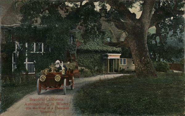 Old photo of people in an open Model T car. The are on a driveway in front of a house covered with vines. Beside the driveway the grass is green. 