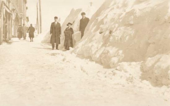 Old photo of two men and a woman standing beside a snow drift that is higher than their heads.