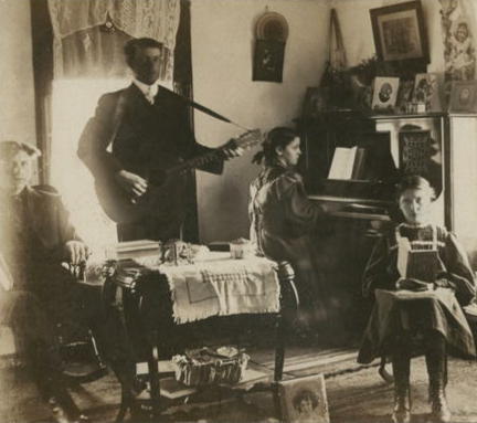 Old photo of family in parlor. Young girl plays piano. Beside her man stands playing guitar. a teen boy and young girl sit at a nearby table. 