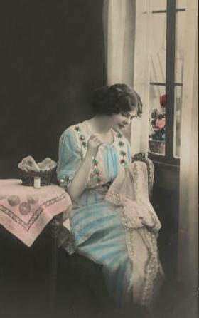 Old photo from about 1915 of a young woman hand-sewing. She is seated on a chair beside a window. Beside her is a table with a small sewing basket and a spool of thread.