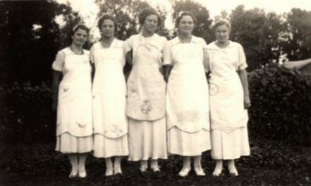 Black and white photo of five ladies standing arm-in-arm in a row. Each is wearing an apron.