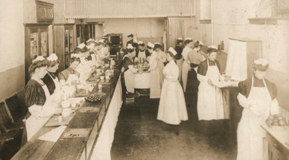 Photo from the early 1900s of a large classroom full of cooking students and teachers. The students are wearing caps and full aprons. The teachers wear caps and are dressed in white. Some students are preparing vegetables, others are setting a table, while others are at different stages of the cooking process. 
