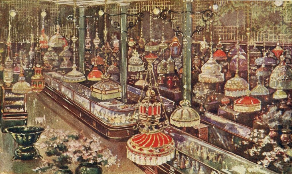 Postcard showing a variety of lanterns, lamps, and chandeliers hanging from the ceiling. Beneath are display cases with smaller items for sale.