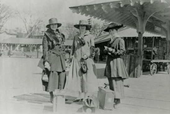 Black and white photo of three women in coats and hats, standing on a train platform with their suitcases on the ground beside them.