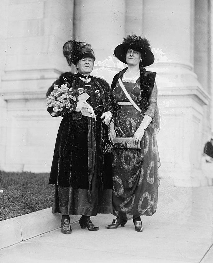 Old photo of two women standing outside on a sidewalk in front of a large building. Both women wear dresses, hats, and gloves commensurate with the early 1920s. Mrs. Lothrop carries a large bouquet of flowers adorned with a large bow.
