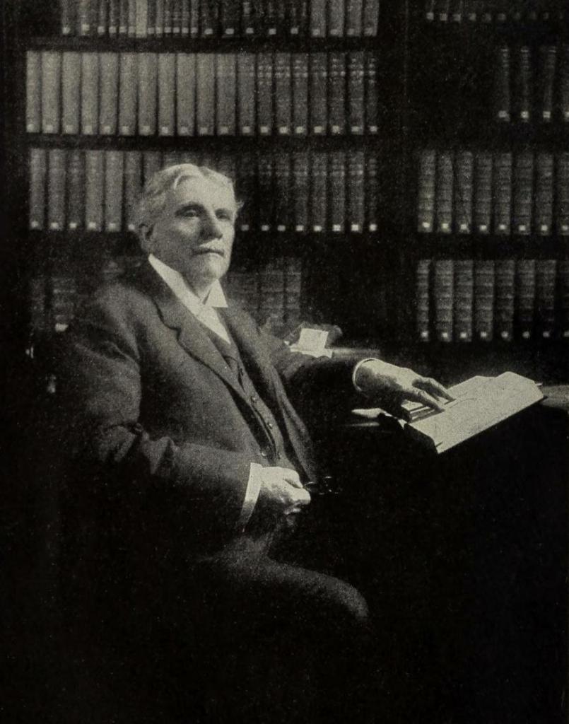 Black and White photo of Cyrus Ingerson Scofield in his later years. His hair is white and he is dressed in a three-piece suit. He is seated at a table.  In one hand he holds a pair of glasses. His other hand points to a passage of text in a Bible open on the table before him. Behind him are bookcases lined with books. 