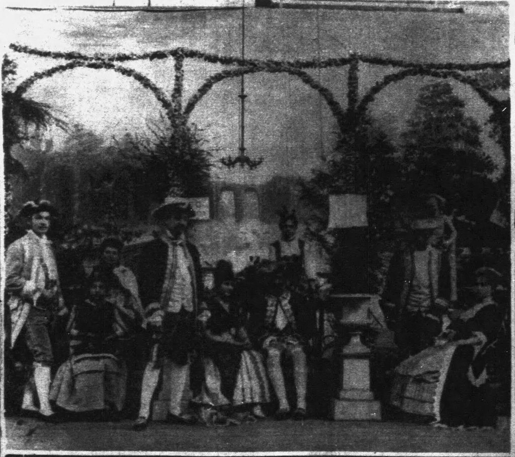 Old black and white photo showing a group of people dressed in costumes from the 1770s. Some men are standing; others are seated beside women. There is a statuary urn with a plant between the chairs. Behind them is a background painting of a garden scene with more statues.