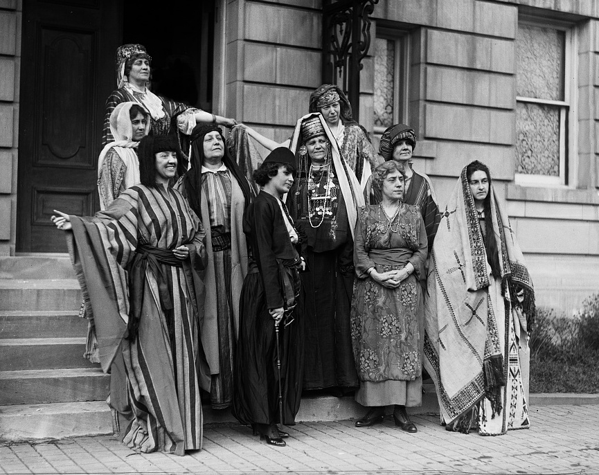 Old photo of ten women standing on the steps of a building. Each woman is dressed in a different costume, such as Native American, Arabian, Western, etc.