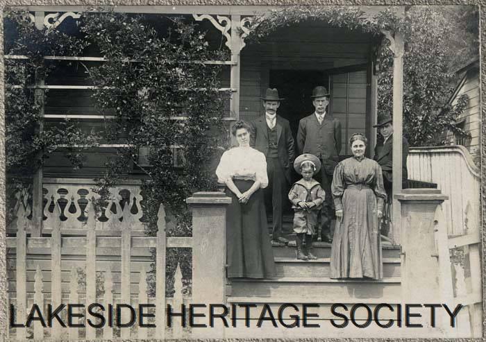 Old photo about 1899 of a family posing on the steps of their cottage. The porch has numerous flowering vines that weave in and out of gingerbread trims. On the steps stand two women and a little boy in a sailor suit. Behind them, two men stand and one man sits; each wears a full suit and hat. 