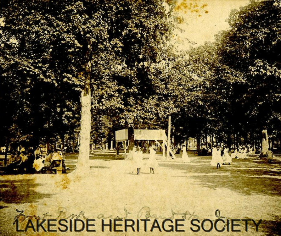 Old sepia photo about 1910 of an open park area with children playing and standing in groups. Behind them is an elevated band stand. Behind the band stand and to the left is a grove of mature trees. People sit in groups in the shade or walk under the trees.