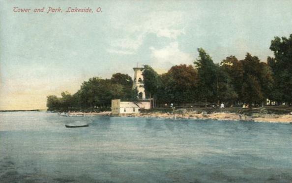 Photo of a promontory point of land on the shore of the lake. a short pier extends from the land out onto the water. Beside the pier is a white building with a tower. To the right along the shore line is a grove of mature trees and a park. 