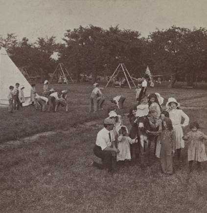 Old photo of a man wearing a cap, white shirt and tie, kneels beside a group of children who are crowded around a small donkey. Behind them a group of children play a game on the grass beside a tepee. In the background children play on swing sets.