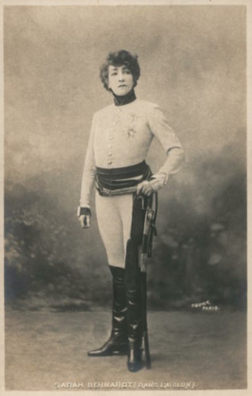 Photo of Bernhardt wearing a pseudo-military uniform of high-collared tunic with long sleeves, and form-fitting pantaloons tucked into knee-high boots. Around her waist she wears a sash from which is hung a sword in a scabbard.