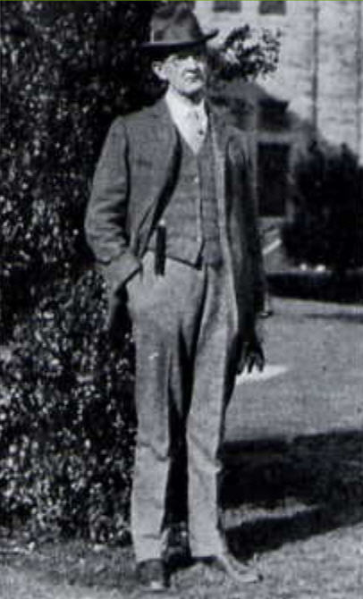 Black and white full-body photo of a gentleman standing out of doors in front of a large stone building. He wears a three-piece suit, a bowler hat and spectacles. One hand is in his trouser pocket; in the other hand he holds a pair of gloves.