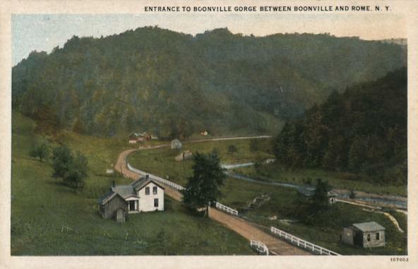 Hand-colored photo (undated) of a farm nestled in a lush green valley between two hills. There is a dirt road bordered by a white picket fence that leads to the white farmhouse. A few outbuildings of different sizes are present.