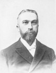 Old black and white portrait photograph from about 1890 of a man in his thirties or forties. His hair is closely cut and he wears a neatly trimmed beard and mustache. He wears a dark colored suit with a white shirt and white tie. 