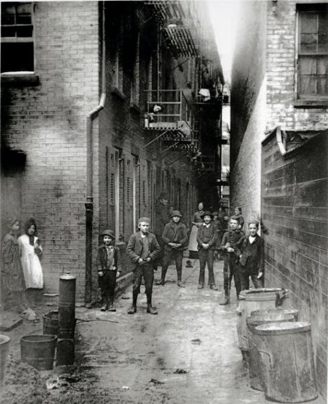 Black and white photo of a narrow alley between two multi-story brick buildings. In the foreground are trash cans and debris, a water pump and buckets. A group of six boys stand in the center of the photo. Behind them are two women wearing long dresses and aprons  from the time period, as well as more children. On the left side of the photo are two young girls, wearing short dresses that appear to be patched.