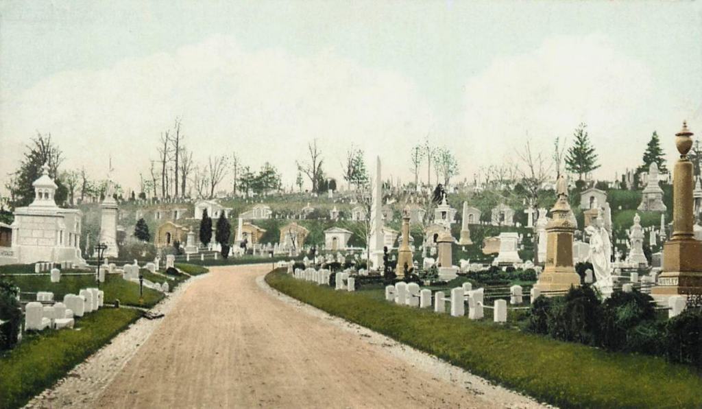 Old photo showing a narrow dirt road. ON either side are small headstones and large statuary monuments marking graves. 