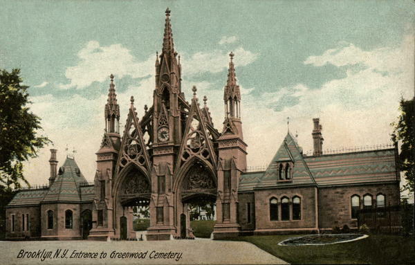 Photo of an elaborate Gothic architecture with pointed arches, intricate stonework, and multiple spires topped with decorative finials. The central structure has large arched openings that serve as gateways, flanked by smaller architectural elements. The building extends to both sides of the entrance with additional Gothic-style structures featuring steep roof lines, dormer windows, and tall chimneys.