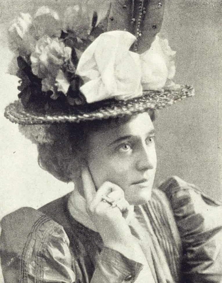 Black and white photo of a woman wearing a dress from the 1890s. On her head is a flat brimmed hat; the crown of the hat is covered with flowers, gathered ribbon, and a single large bird feather in the front. The feather stands straight up on its stem and is adorned with beads.