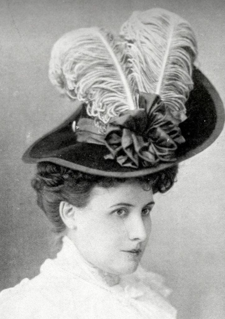 Black and white photo of a woman wearing a short-brimmed hat. A wide ribbon circles the crown of the had and is arranged in multiple loops in front. The stems of two long ostrich feathers are tucked into the ribbon arrangement and the feathers drape back cross the crown of the hat.