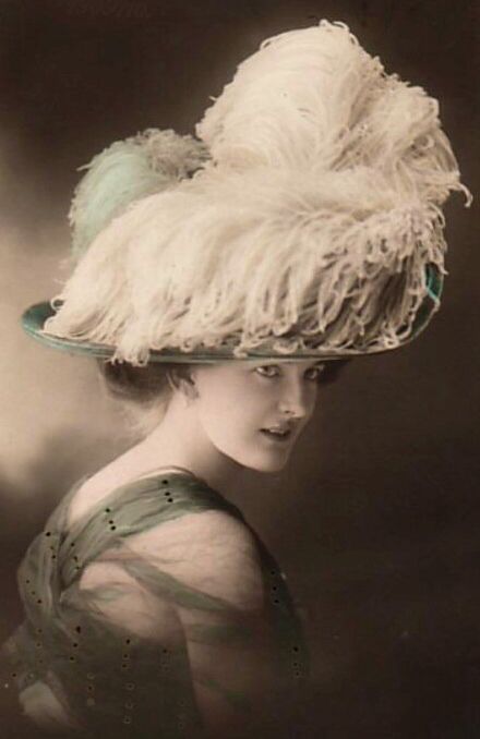 Hand-colored photo of a woman wearing a hat with a wide brim of 12 to 14 inches deep. The crown of the hat is covered in ostrich feathers that are piled high, with some parts of the feathers spilling over the brim of the hat.