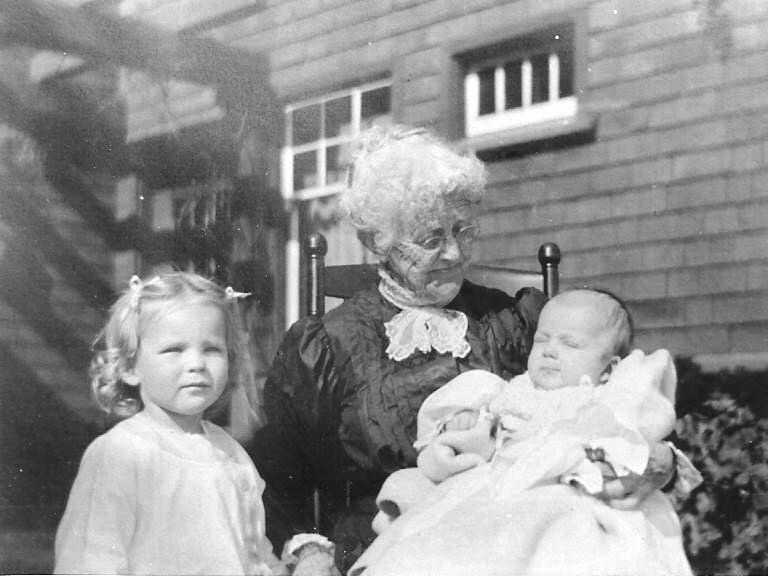 Black and white photo of an elderly woman in her 80s, seated in a chair. On her lap she holds a baby and beside her stands a little girl about 3 years old. 