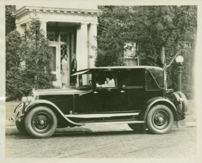 Black and white photo of a side-view of an old automobile with four doors and a woman seated in the driver's seat.