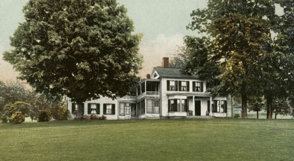 Color photo of a sprawling two-story home set amid mature green trees, surrounded by green lawns. The house is white with a dark green shingled roof and window shutters. A small covered porch marks the entry door. Covered verandas on the first and second stories offer shade. 