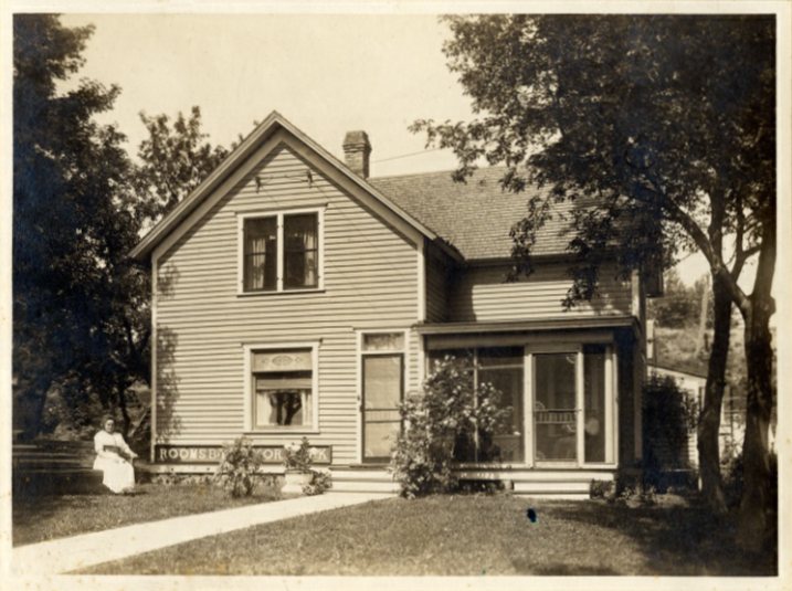 Black and white photo from about 1910 showing a neat two-story house with clapboard siding and a small screened front porch. A woman sits alone in a chair to the left of the house near a shade tree.