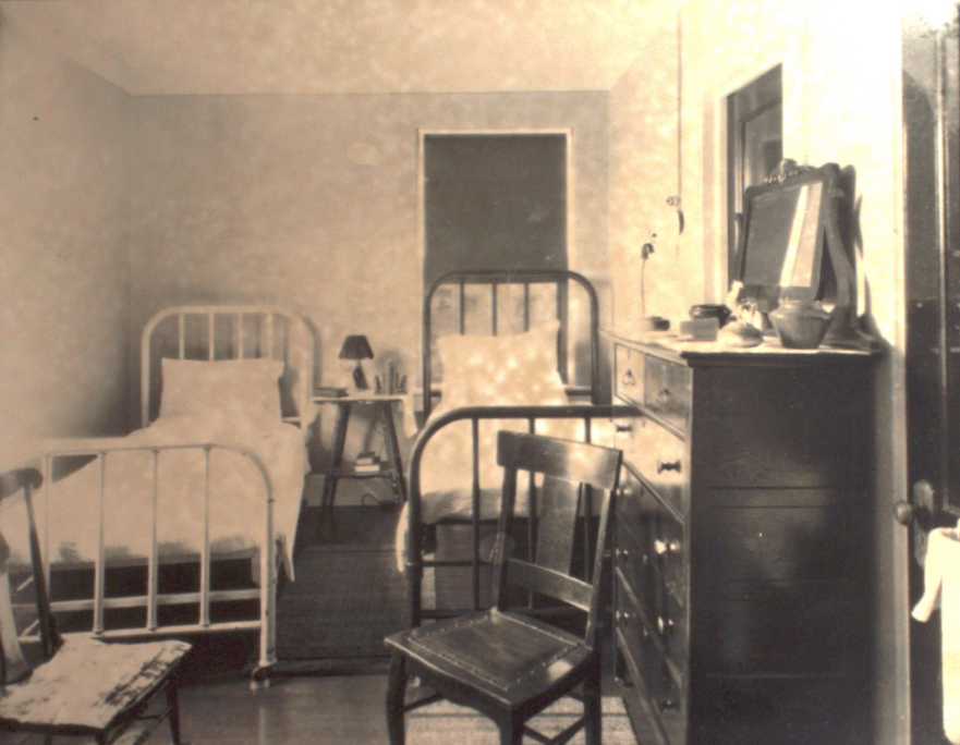 Black and white photo of a small room containing two twin beds with iron frames. At the foot of each bed is a wooden chair. A tall chest of drawers stands against a wall between two doors. On top of the chest are a mirror and small toiletry items and jars. In the foreground you can see what appears to be the edges of towels hanging from a rod. One small window is covered by a dark shade.