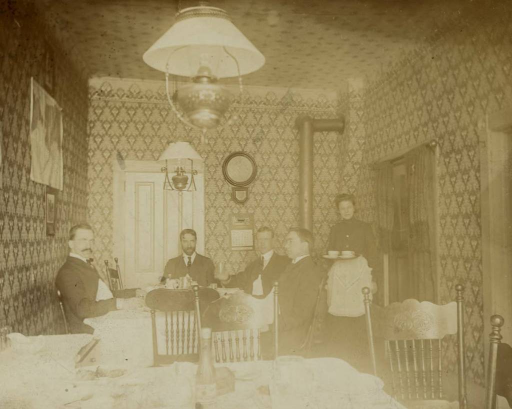 Old black and white photo of a small dining room with 2 gas lamps hanging from the ceiling. Four men sit at a table covered in a white table cloth as a woman wearing an apron carries a tray toward them. In the foreground is an empty table and chairs set with linens, dishes and cutlery. The walls and ceiling are covered with patterned wallpaper. In the back corner of the room you can see the exhaust pipes for a small wood-burning stove.