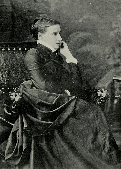 Black and white photo of Elizabeth Stuart Phelps-Ward about 18905. Seated in a chair, her face in profile. Her hair is drawn back and arranged in a modest chignon. She wears a dark gown with a white ruffle trimming the cuffs of her long sleeves and a high color.